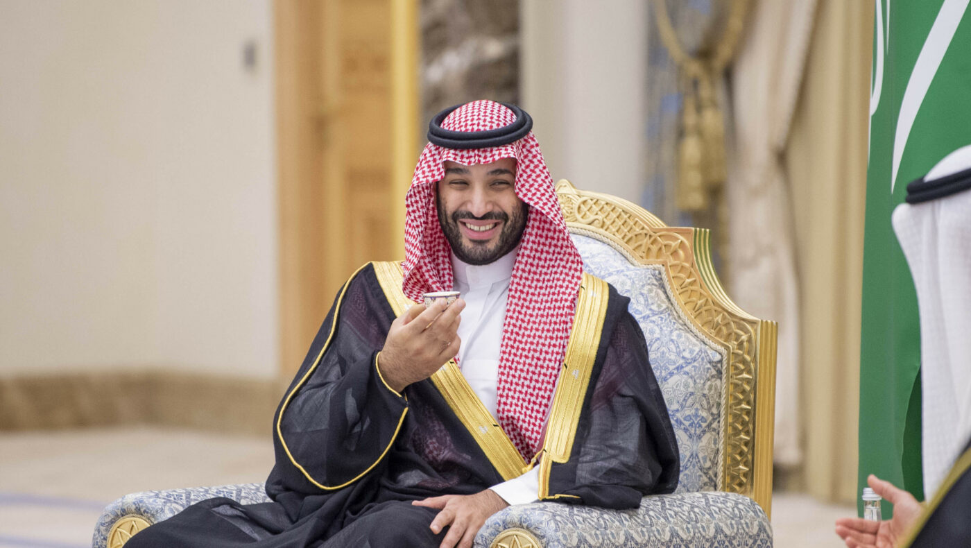 Mohammed bin Salman lächelt und erhebt eine Hand während er auf einem Stuhl sitzt
