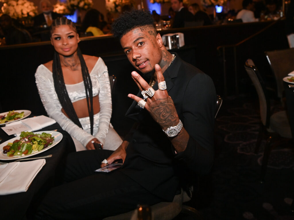 Rapper Blueface, schwar angezogen hält seine tättowierte Hand mit großen Ringen und einer auffälligen Uhr in die Kamer. Dahinter sitzt Chrisean Rock in weißem bauchfreiem Outfit