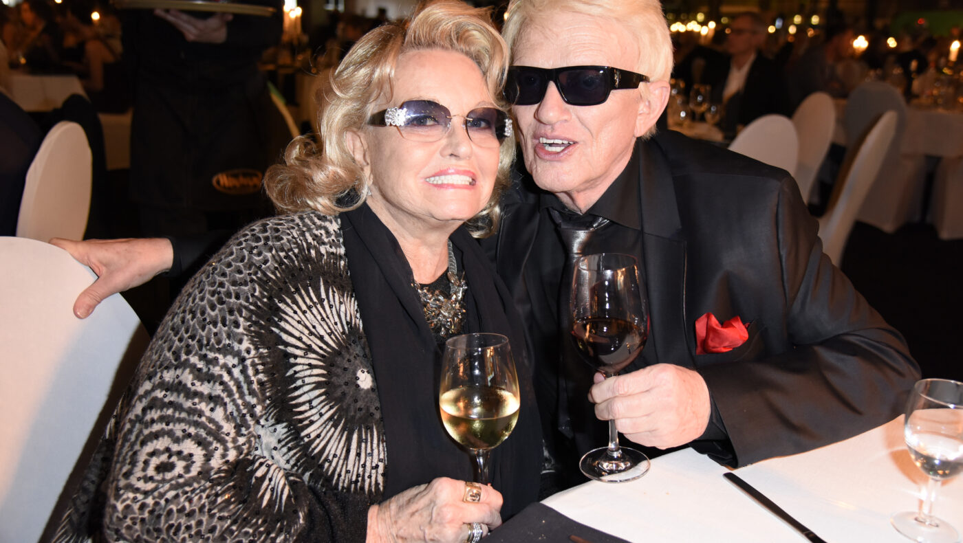 Hannelore und Heino sitzen an einem Tisch. Hannelore hält Glas Wein in der Hand.