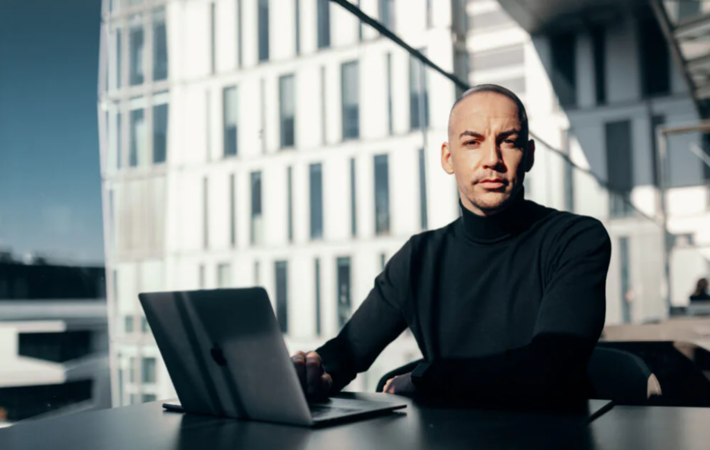 Erfahrungen Poosch Consulting GmbH Phil Poosch sitzt mit schwarzem Pullover an einem PC neben einer riesigen Glasfront durch die ein Hochhaus zu sehen ist