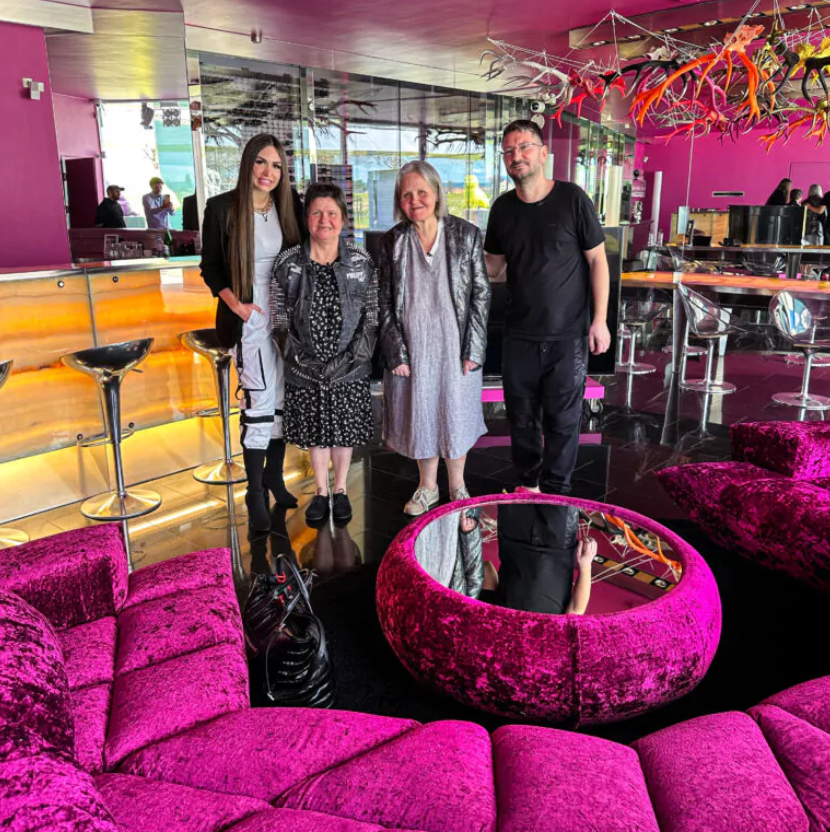 Walter und Susi Temmer stehen mit zwei Damen in ihrer Villa vor einem pinken Plüschsofa. Dahinter bunte Hirschgeweihe und Barhocker