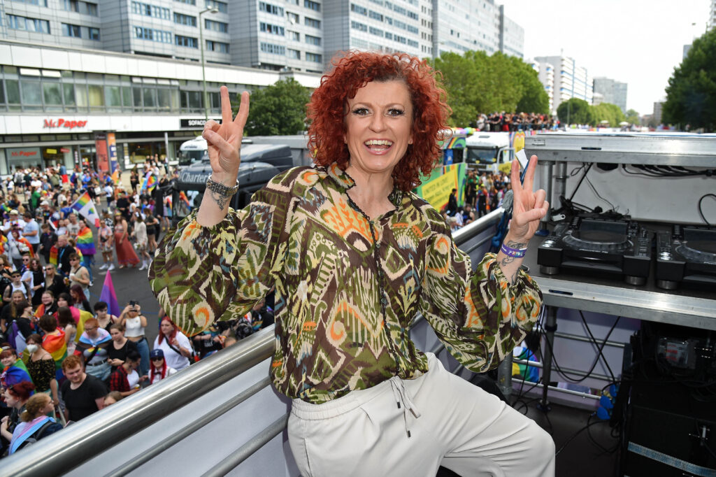 Lucy Diakovska mit einer bunten Bluse und weißer Hose streckt beide Hände mit dem Peace-Zeichen in die Höhe und lächelt in die Kamera. Hinter ihr viele Menschen mit Regenbogenfahnen auf der Straße