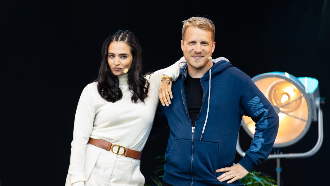 Amira und Oliver Pocher. Sie trägt ein weißes Outfit und stützt sich lässig auf ihren Mann, der eine blaue Sportjacke trägt