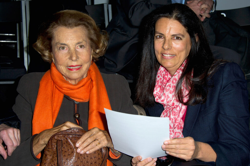 10 reichsten Frauen der Welt Francoise Bettencourt-Meyers mit blauem Sakko und rot-weißem Schal hält ein Blatt Papier in ihren Händen. Neben ihr eine elegante Dame mit dunklem Kostum und orangem Schal