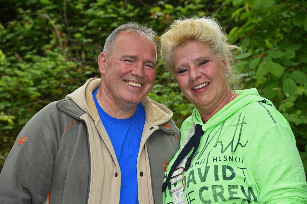 Silvia Wollny mit ihrem Partner. Sie trägt ein grünes T-Shirt, er eine Jacke und ein blaues T-Shirt. Beide lächeln in die Kamera