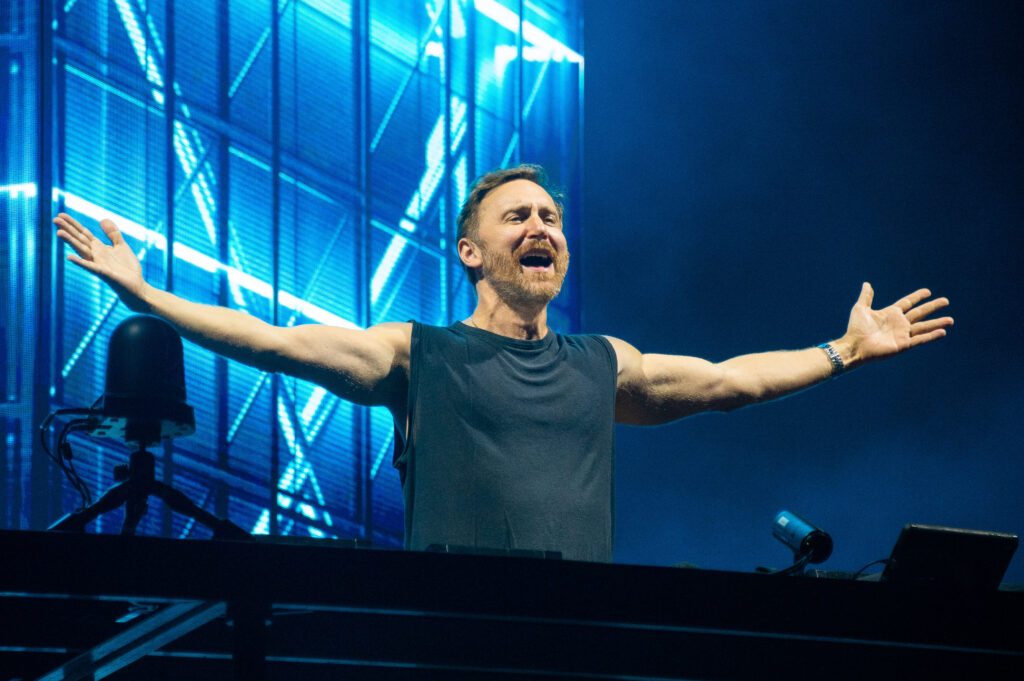 David Guetta steht an einem Mischpult und breitet die Arme weit aus. Hinter ihm ein blauer Hintergrund mit Schweinwerfern bestrahlt