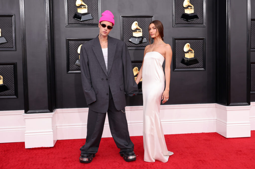 Justin und Hailey Bieber stehen vor einer schwarzen Wand die goldene Plattenspieler zeigt; Hailey trägt ein bodenlanges weißes Kleid und Justin einen schwarzen riesigen Anzug, eine schwarze Sonnenbrille sowie eine pinke Mütze