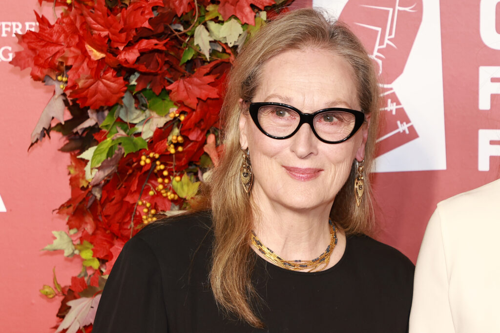 Meryl Streep vor einem Plakat mit dunkelroten Blättern. Sie trägt eine schwarze Brille und ein schwarzes Oberteil