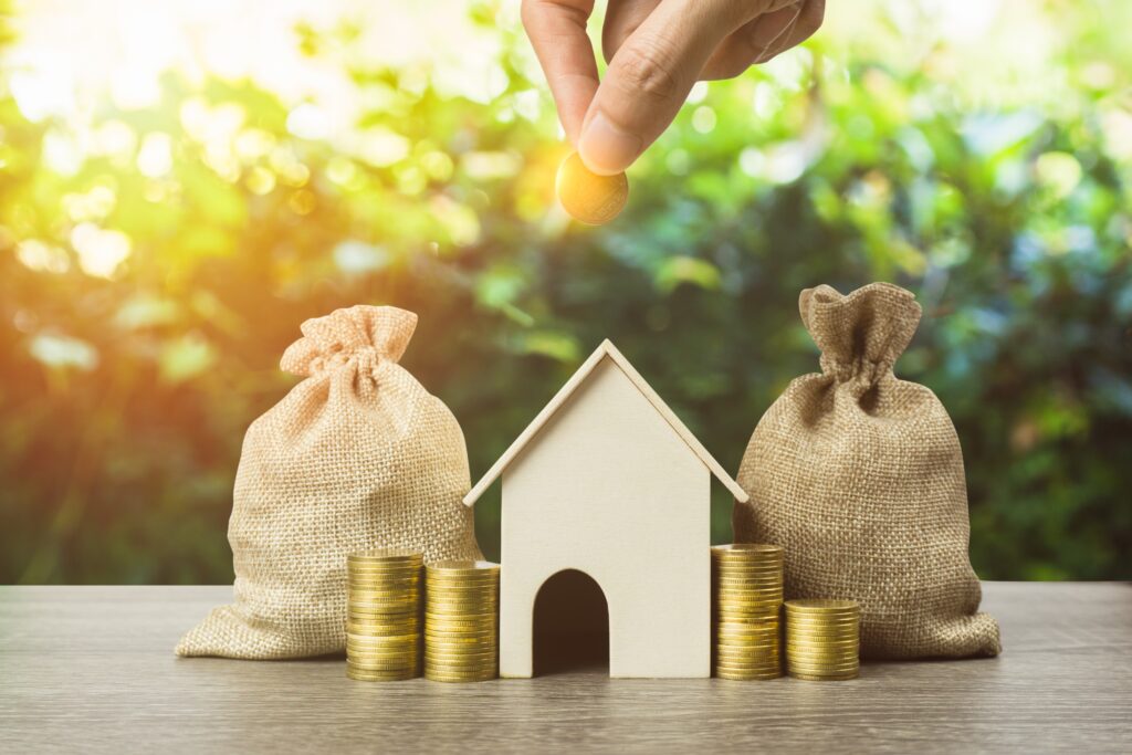 Goldgrüner unscharfer Hintergrund. Davor zwei Jutesäcke. in der Mitte ein Haus und neben dem Haus jeweils zwei Stapel mit Goldmünzen auf einer Holzplatte