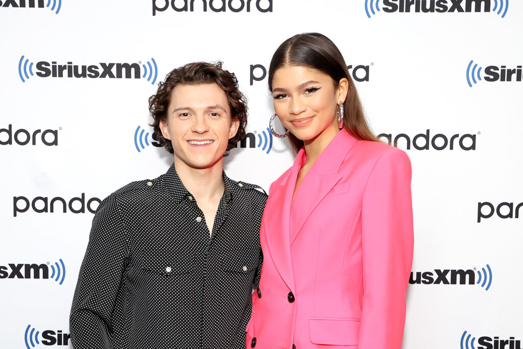 Zendaya und Tom Holland stehen gemeinsam vor einem Plakat. Er trägt ein schwarzes Oberteil und sie eine pinkfärbiges Sakko