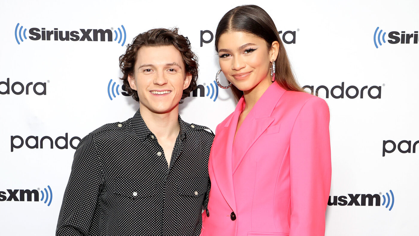 Zendaya und Tom Holland stehen gemeinsam vor einem Plakat. Er trägt ein schwarzes Oberteil und sie eine pinkfärbiges Sakko