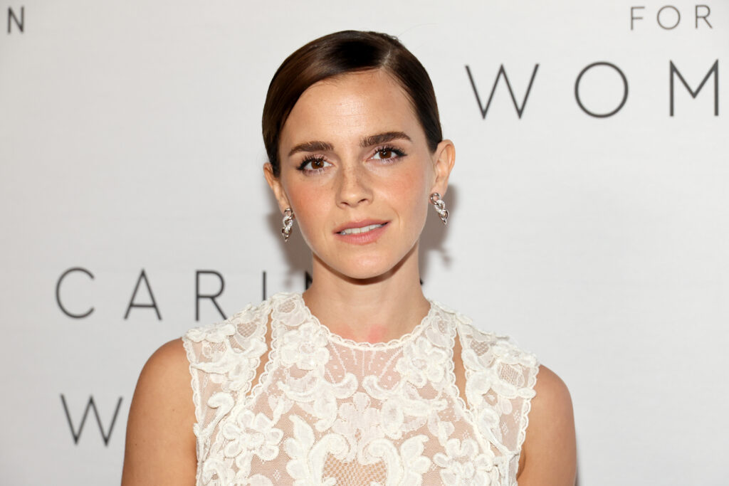 Emma Watson mit einem weißen Spitzenoberteil steht vor einem Plakat mit der Aufschrift: Caring vor Women