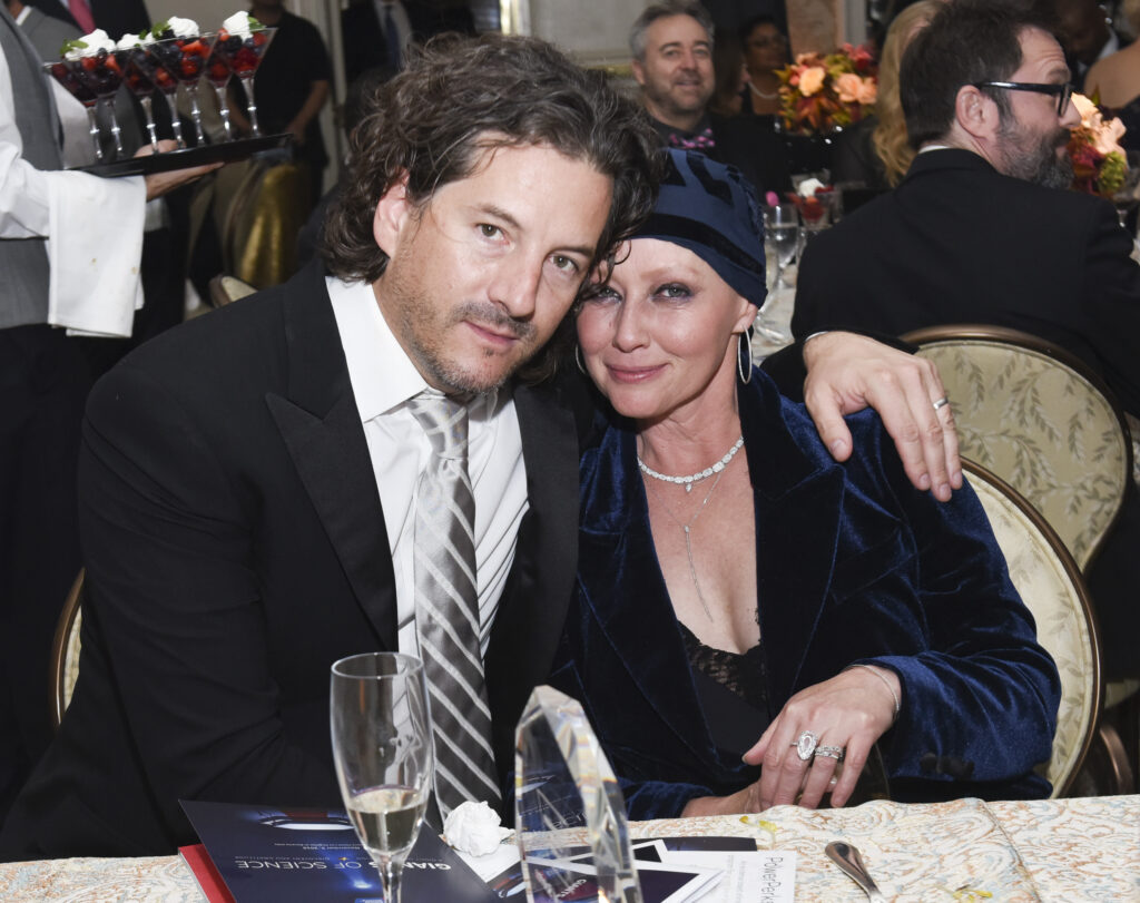 Shannen Doherty Shannen Doherty mit Kopfbedeckung und elegantem Kleid sitzt mit einem Mann in Anzug an einem Tisch