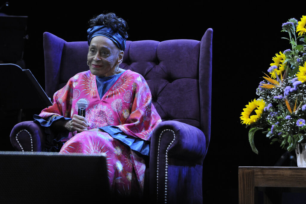 Omara Portuondo mit buntem rosa Kleid sitzt auf einem Sofa-Sessel
