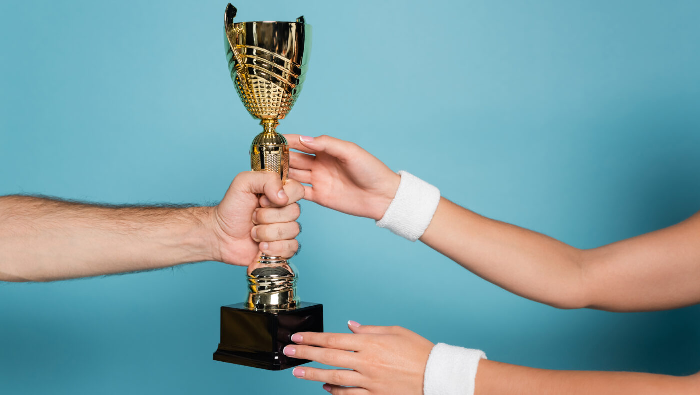goldener Pokal der von einer Hand gehalten wird. Zwei weitere Hände greifen danach