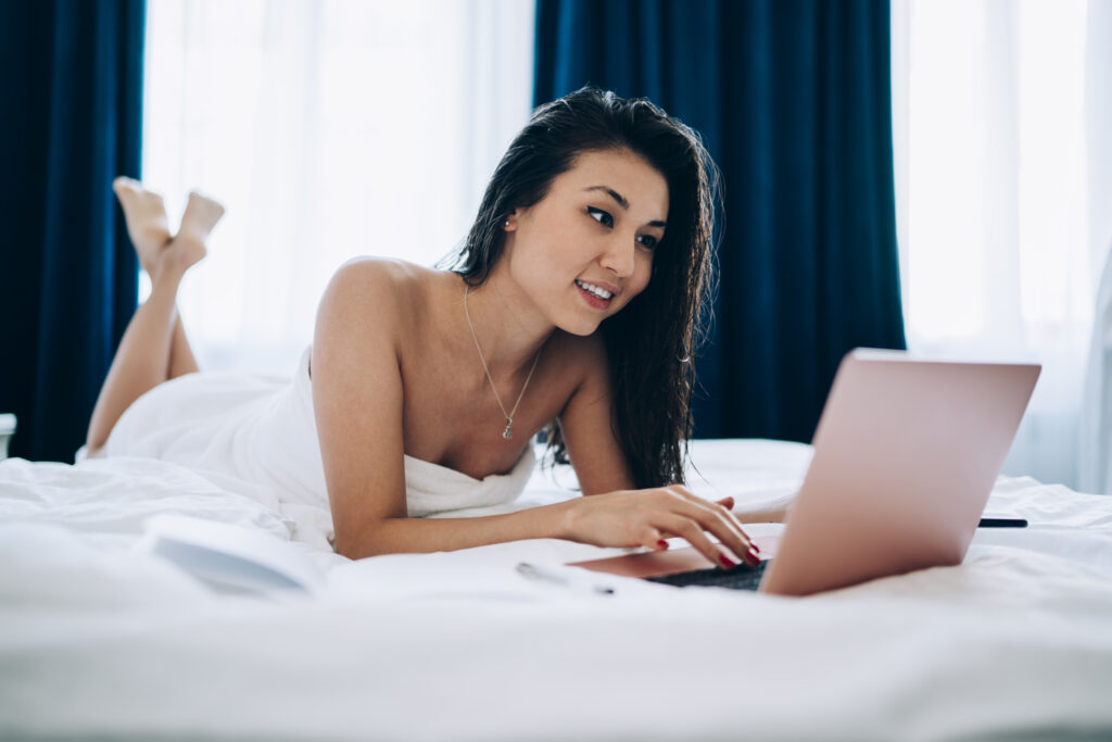 Frau liegt mit einem Handtuch eingewickelt auf einem Bett und hat ein Notebook vor sich