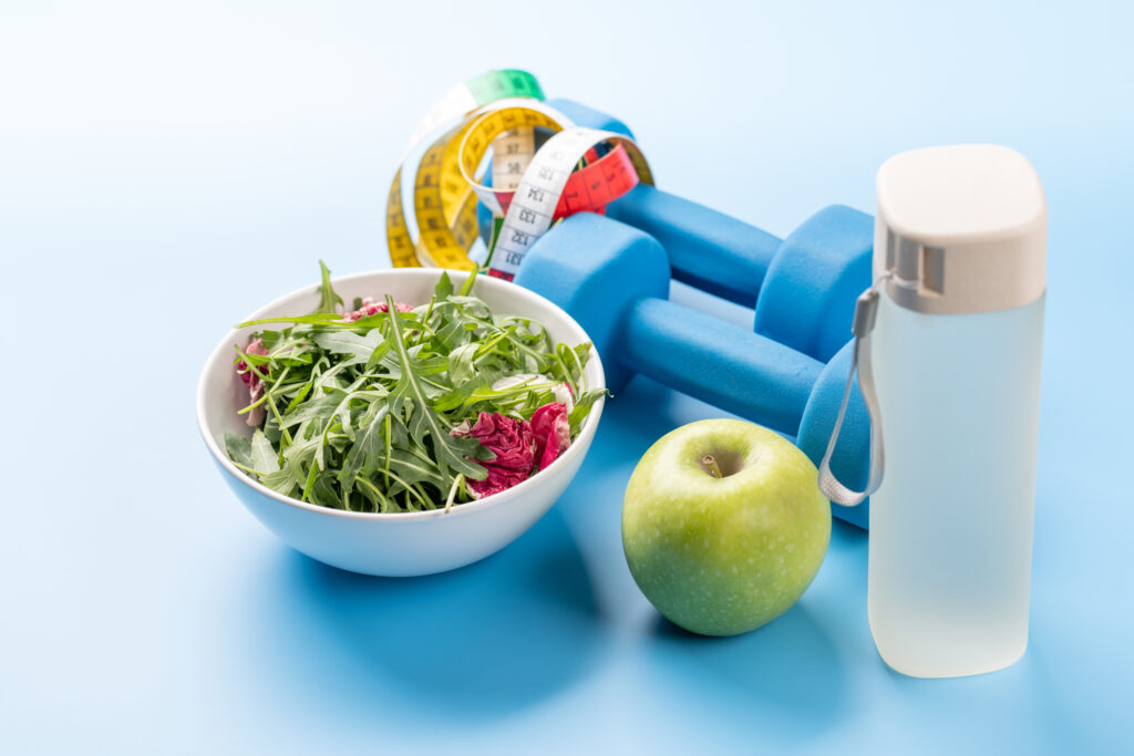 roher Salat, ein Apfel, eine Wasserflasche, zwei blaue Gewichtshandeln und ein Maßband