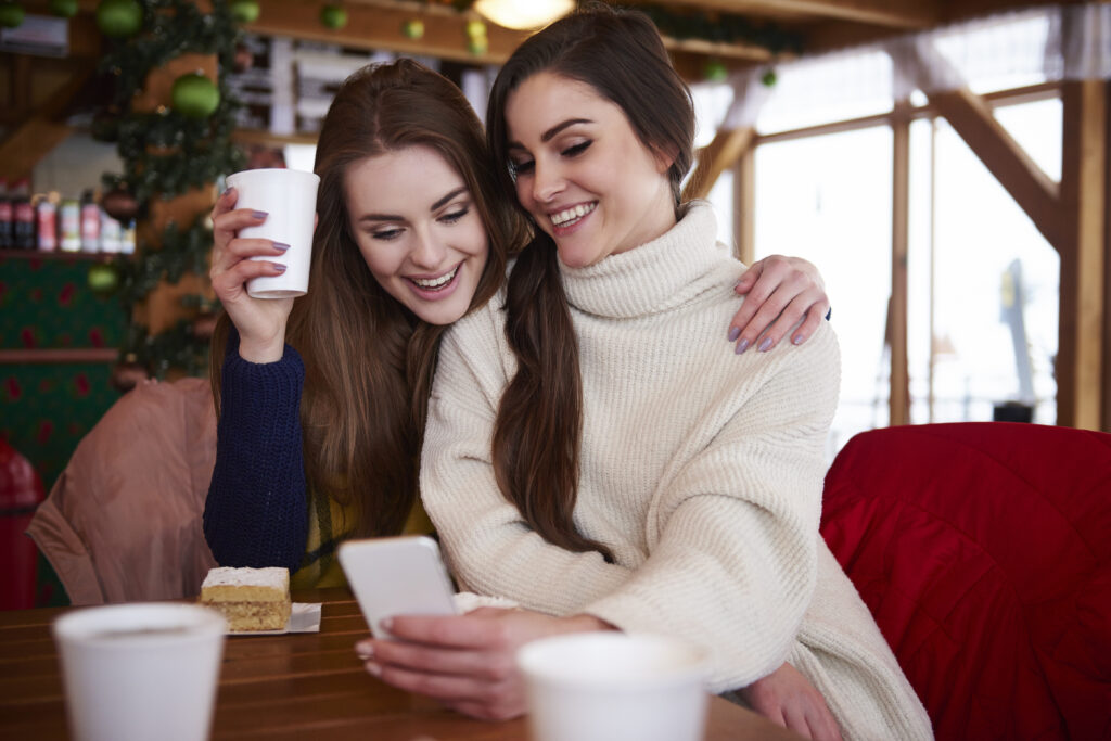 Zwei Frauen schauen auf ein Handy. Die eine hält einen Kaffeebecher, die andere das Handy. Sie sitzen an einem Tisch auf dem ein Kuchen steht