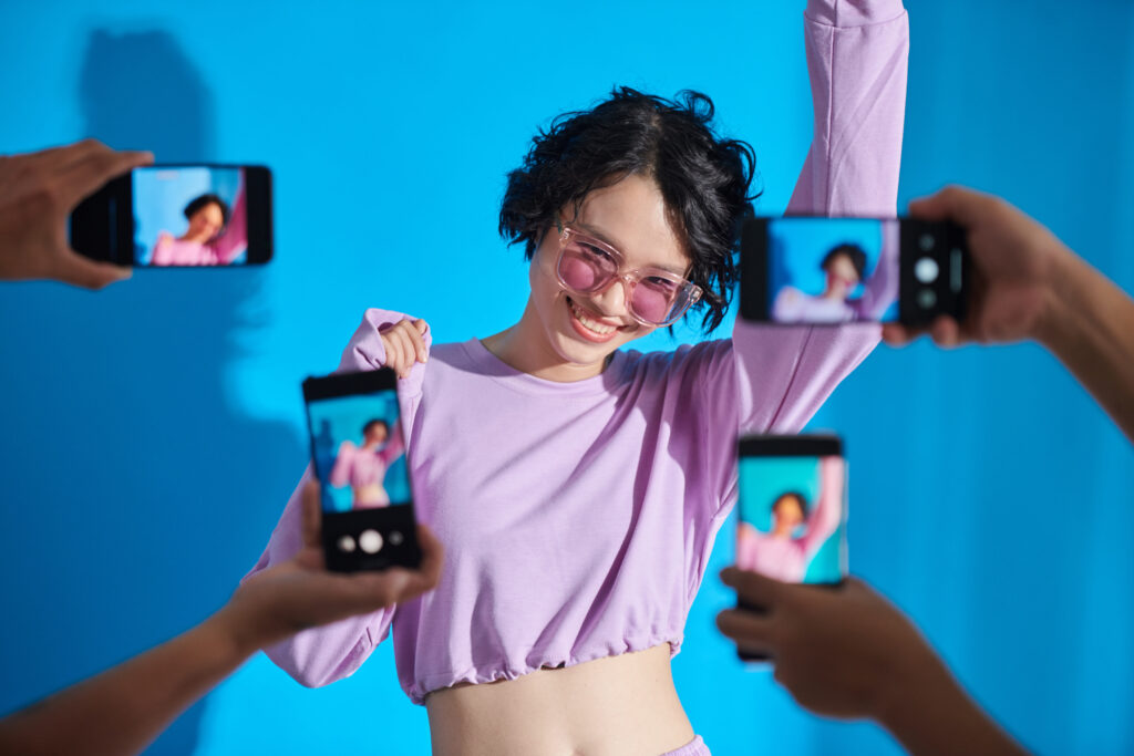 Frau mit pinkem Oberteil und pinker Sonnenbrille hebt eine Hand in die Höhe. Vor ihr vier Handys die sie fotografieren